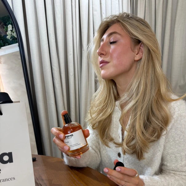 Woman holding a bottle of aroma avenue product in a room with curtains and a mirror.